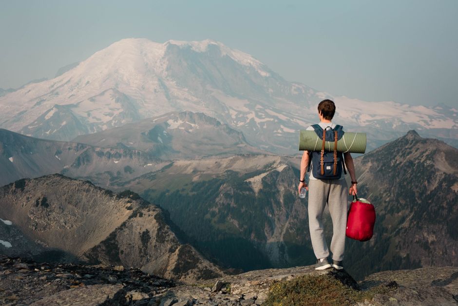 Full body of anonymous male hiker with backpack standing on top of cliff with bag in hand and admiring view of snowy mountain peak